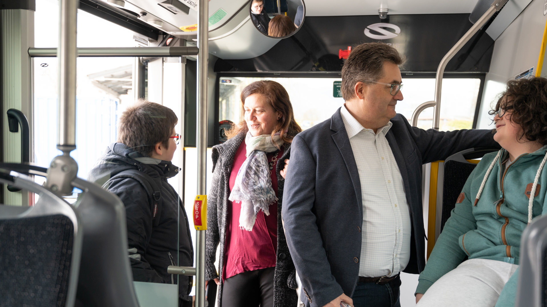 4 passengers, 2 adults and 2 young people, are talking to each other on the bus. One of them is sitting.