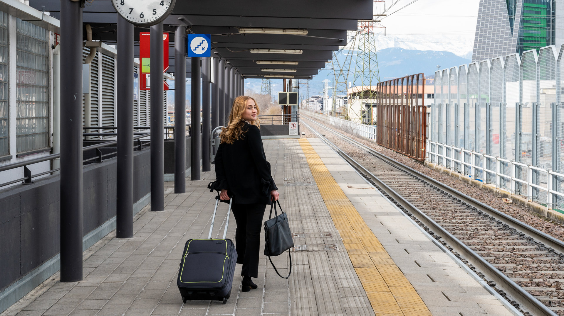 A woman with a bag and a suitcase is walking down the train platform. She is looking towards the rail tracks.